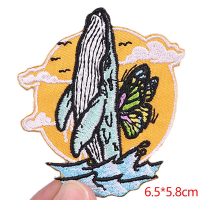 Embroidered patch with whale and butterfly design held by a hand, on a white background.