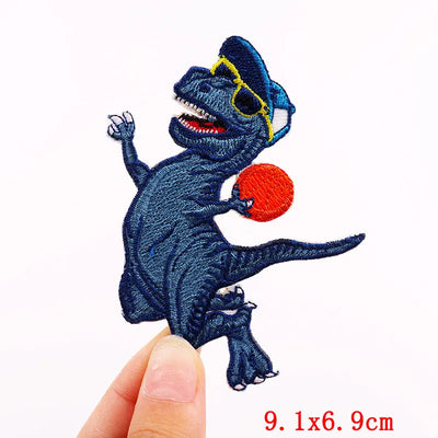 Blue dinosaur patch with sunglasses and a ball, held by a hand on a white background.