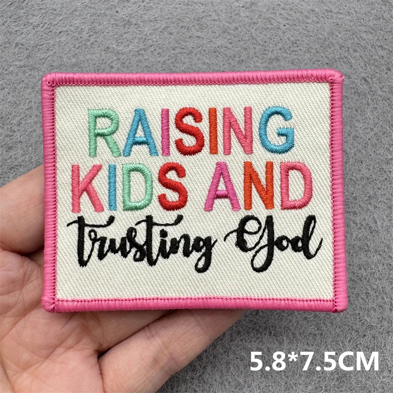 Hand holding a multicolor pink and white embroidered Raising Kids and Trusting God iron-on patch approximately 5.8x7.5cm on a gray background