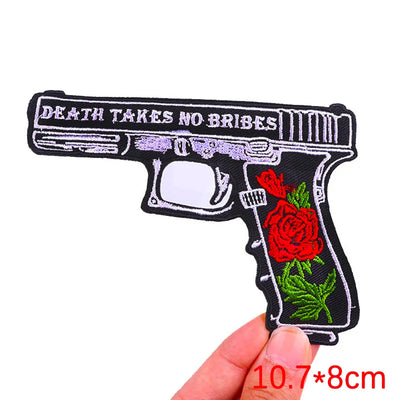 Embroidered patch of a gun with roses and 'Death Takes No Bribes' text, held by a hand.