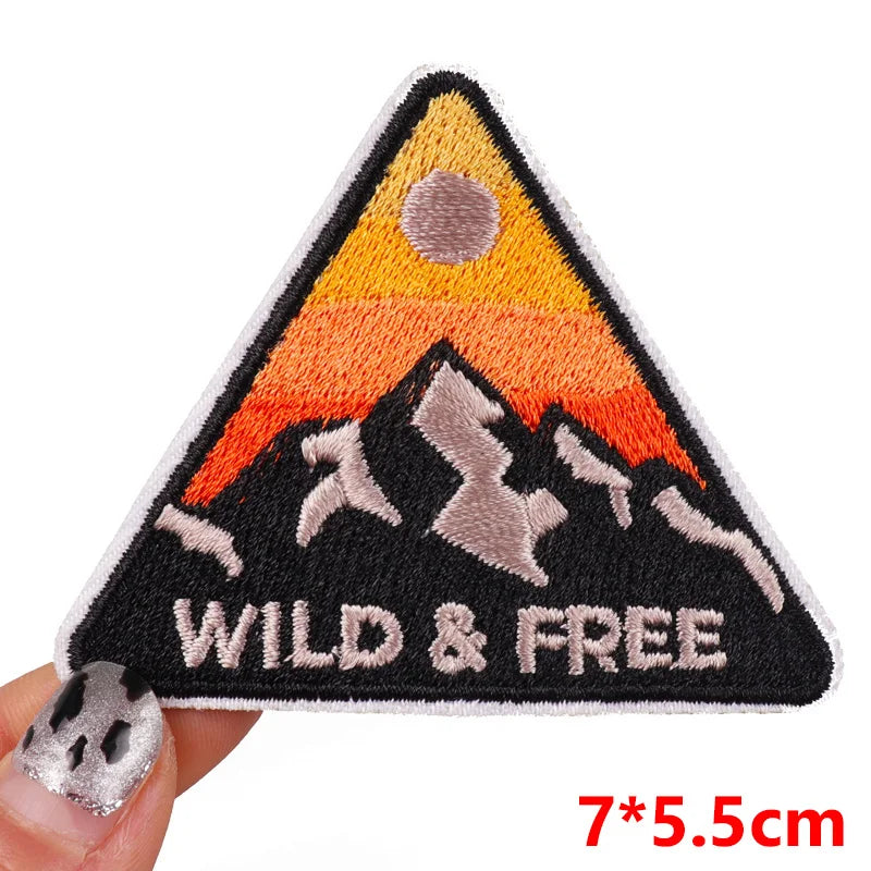 Triangular patch with mountain design and 'Wild & Free' text held by a hand on a white background.