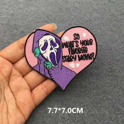 Heart-shaped patch with a ghost face design and text held by a hand on a gray background