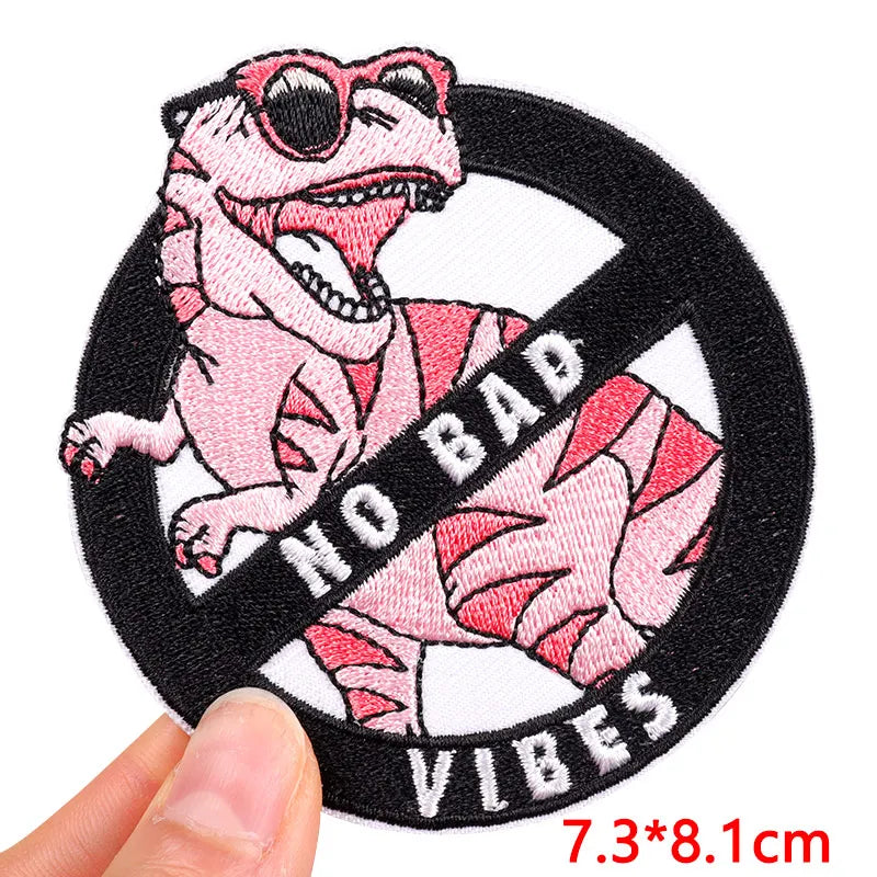 Pink dinosaur patch with sunglasses and 'No Bad Vibes' text held by a hand.