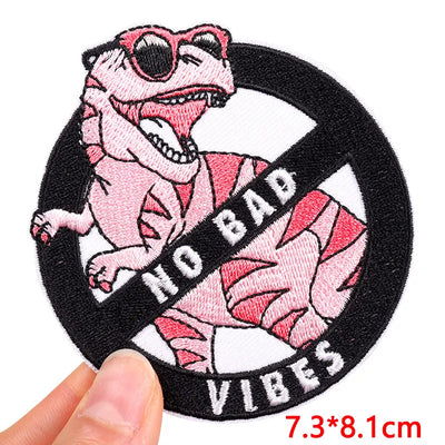 Pink dinosaur patch with sunglasses and 'No Bad Vibes' text held by a hand.