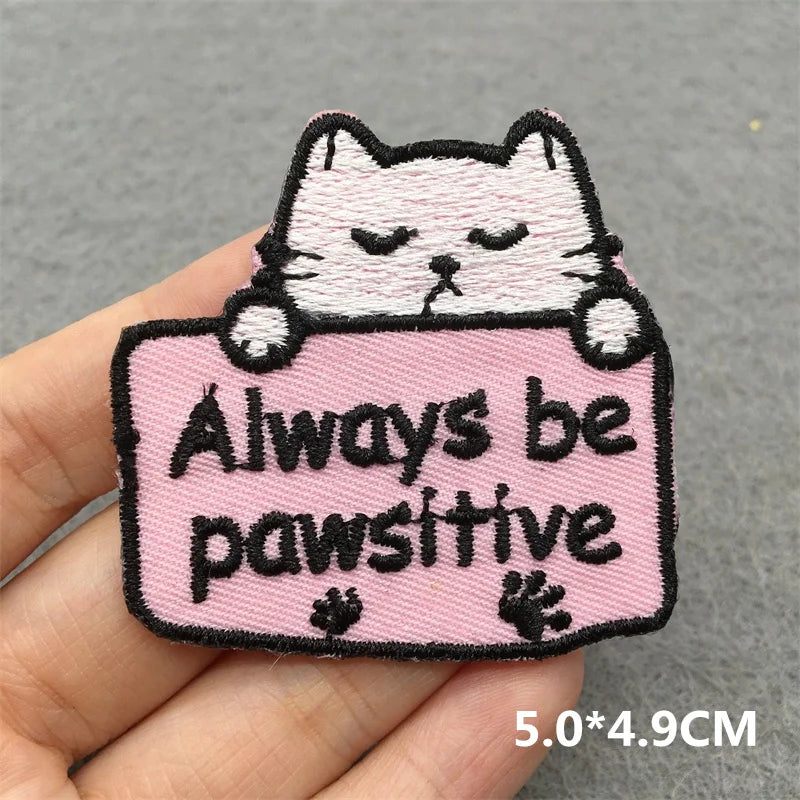 Embroidered patch with a cat design and 'Always be pawsitive' text held by a hand on a gray background.