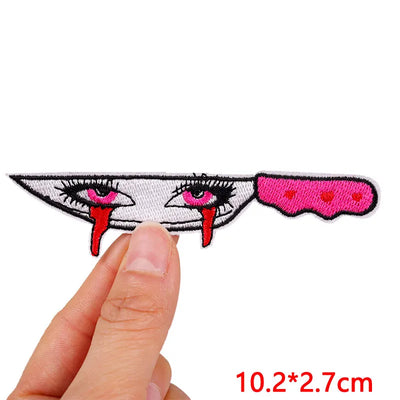 Embroidered patch of a face with red eyes and tongue, held by a finger on a white background.