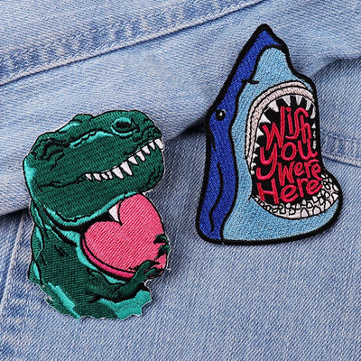 Two patches on a denim background: one dinosaur with a heart, the other shark with text.