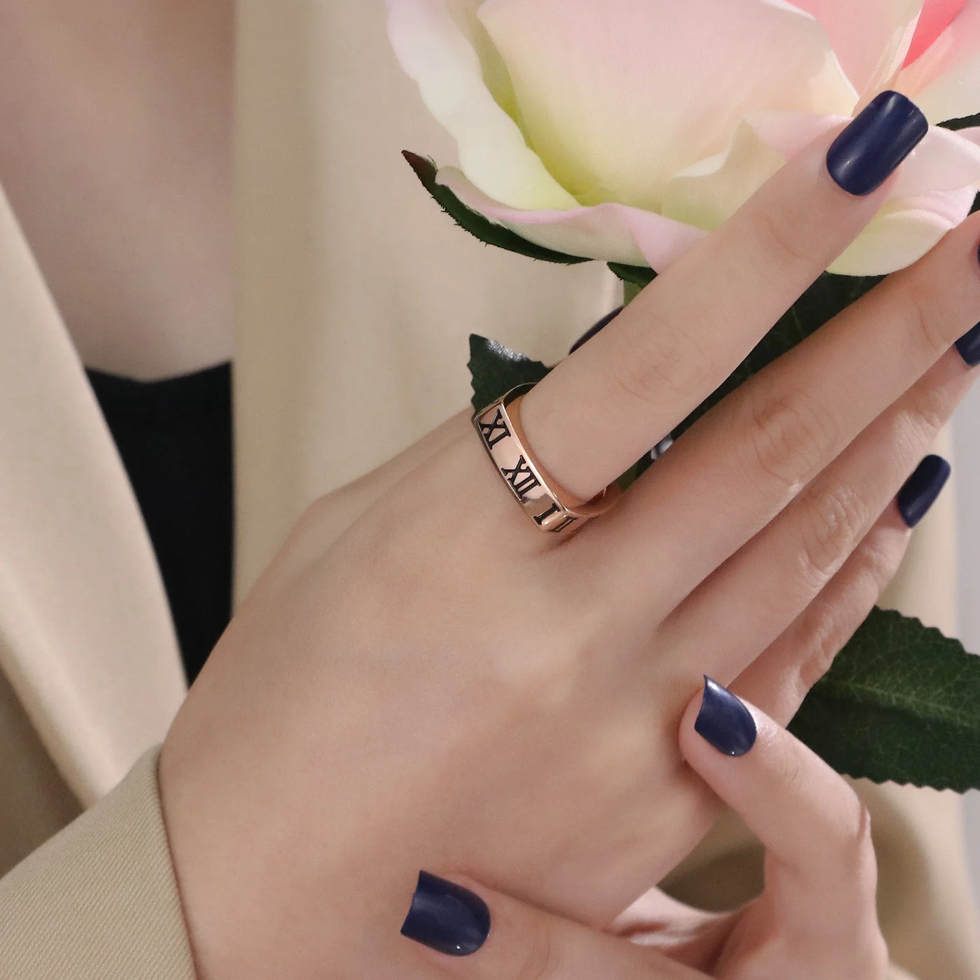 Hand wearing a rose gold ring with Roman numeral design, holding a pink flower.