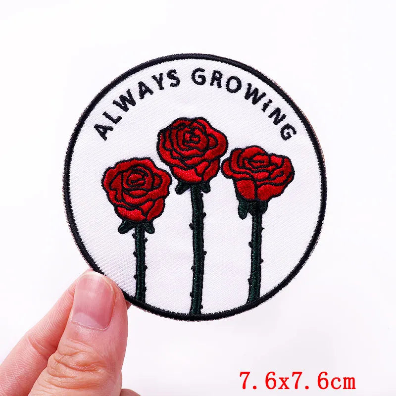 Circular patch with red roses and 'Always Growing' text held by a hand on a white background.