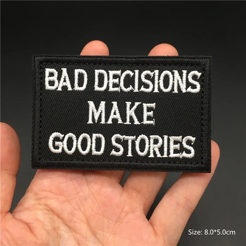 Black patch with white text 'BAD DECISIONS MAKE GOOD STORIES' held in a hand against a dark background.