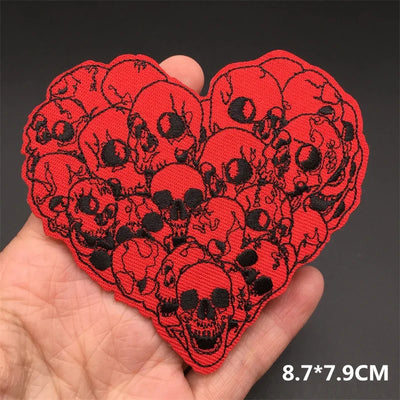 Heart-shaped red patch with black skull designs held in a hand against a dark background.
