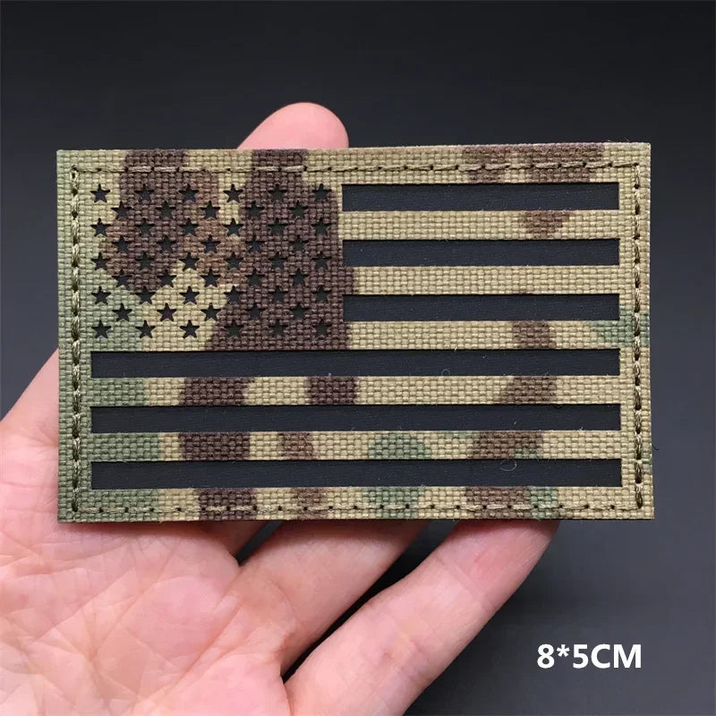 Hand holding a multicam USA flag IR style hook-and-loop tactical badge approximately 8x5cm on a dark background