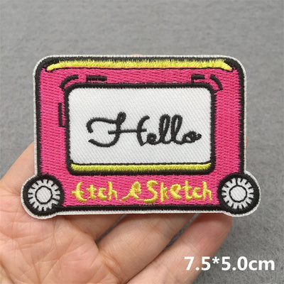 Embroidered patch of a pink etch-a-sketch with 'Hello' text held in hand on a gray background