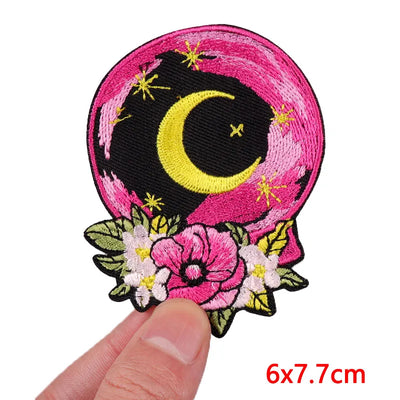 Embroidered patch with a moon and floral design held by a hand, on a white background.