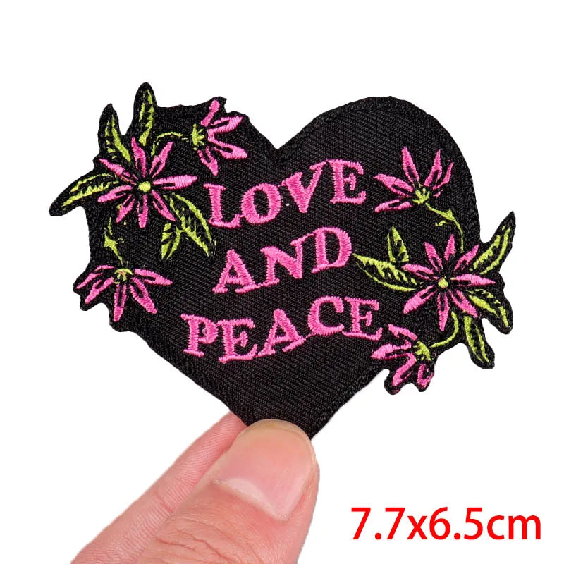 Heart-shaped patch with 'Love and Peace' text and floral designs held by a finger on a white background.