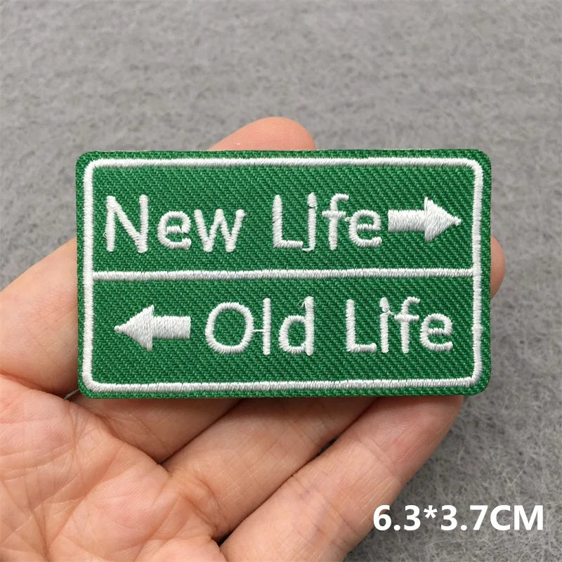 Embroidered patch with 'New Life' and 'Old Life' text held in a hand against a gray background.