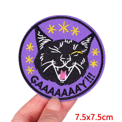 Circular patch with a black cat design and yellow stars on a purple background, held by a hand.