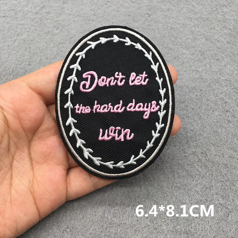 Hand holding a black pink and white embroidered Don’t Let the Hard Days Win iron-on patch approximately 6.4x8.1cm on a gray background