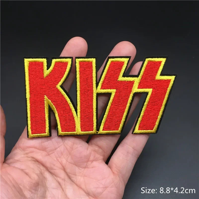 Hand holding a red and yellow Kiss logo embroidered iron-on patch 8.8x4.2cm on a dark background
