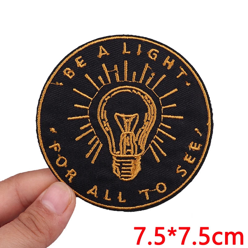 Circular patch with light bulb design and text 'Be a light for all to see' held by a hand.