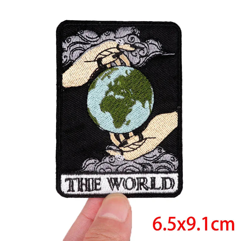 Hand holding a tarot-inspired embroidered iron-on patch featuring hands holding the Earth and the title The World approximately 6.5x9.1cm on a white background