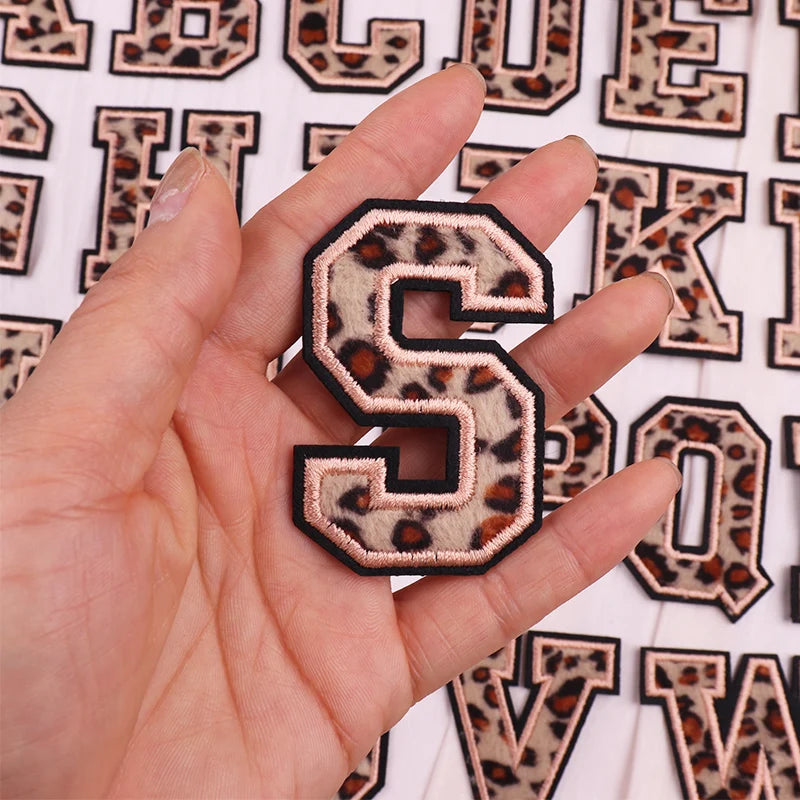 Hand holding a leopard print letter 'S' patch against a background of similar patches.