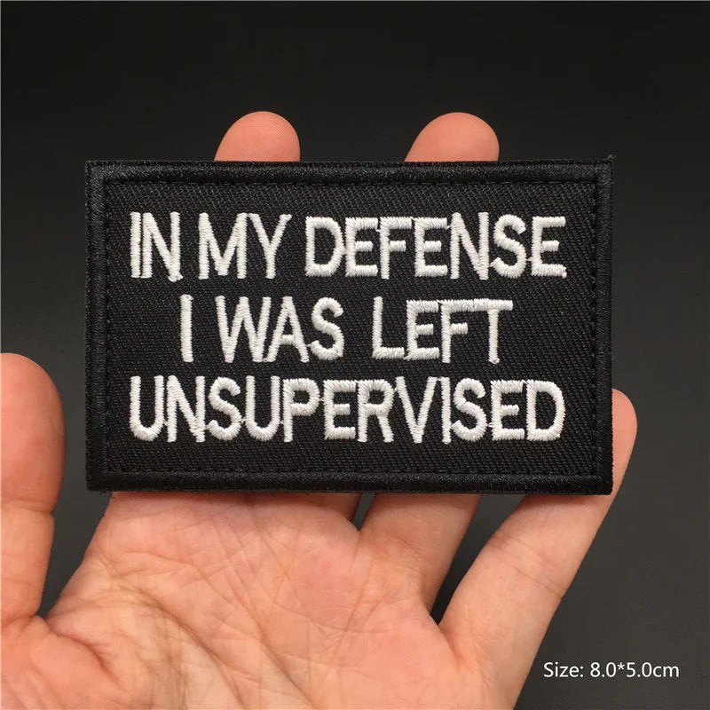 Black patch with white text 'IN MY DEFENSE I WAS LEFT UNSUPERVISED' held in a hand against a dark background.