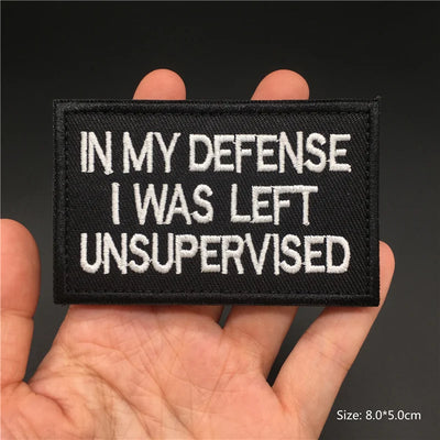 Black patch with white text 'IN MY DEFENSE I WAS LEFT UNSUPERVISED' held in a hand against a dark background.