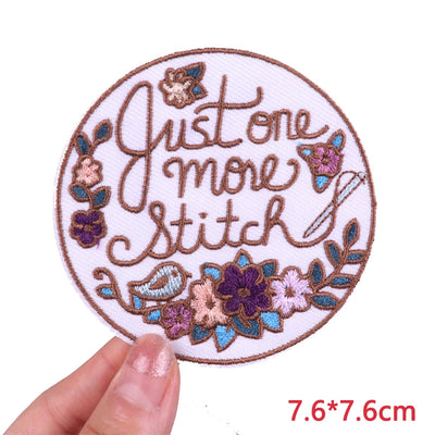 Circular embroidered patch with 'Just one more stitch' text and floral designs, held by a hand.
