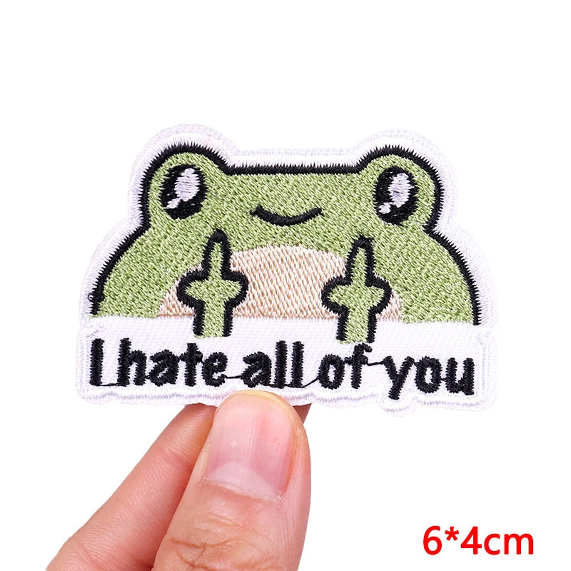 Embroidered patch with a frog design and text 'I hate all of you' held by a hand.