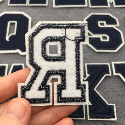 Embroidered patch of a letter 'Q' held over a background with more embroidered letters.