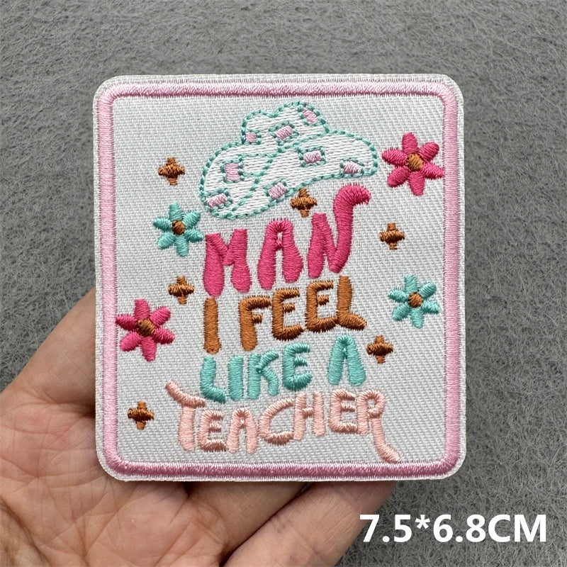 Hand holding a pastel pink turquoise and brown embroidered Man I Feel Like a Teacher iron-on patch with cowgirl hat and floral accents approximately 7.5x6.8cm on a gray background