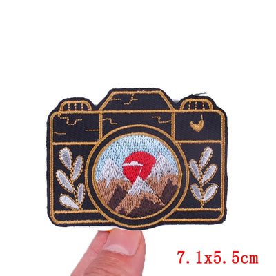 Camera-shaped embroidered patch with a scenic design held by a hand on a white background