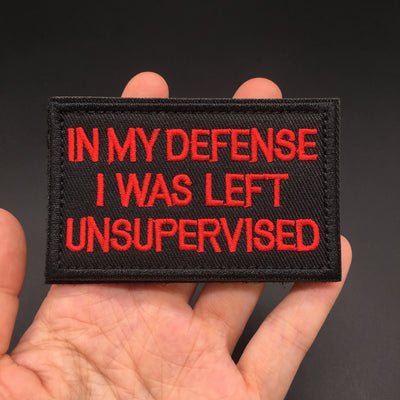 Patch with red text 'IN MY DEFENSE I WAS LEFT UNSUPERVISED' held in a hand against a dark background