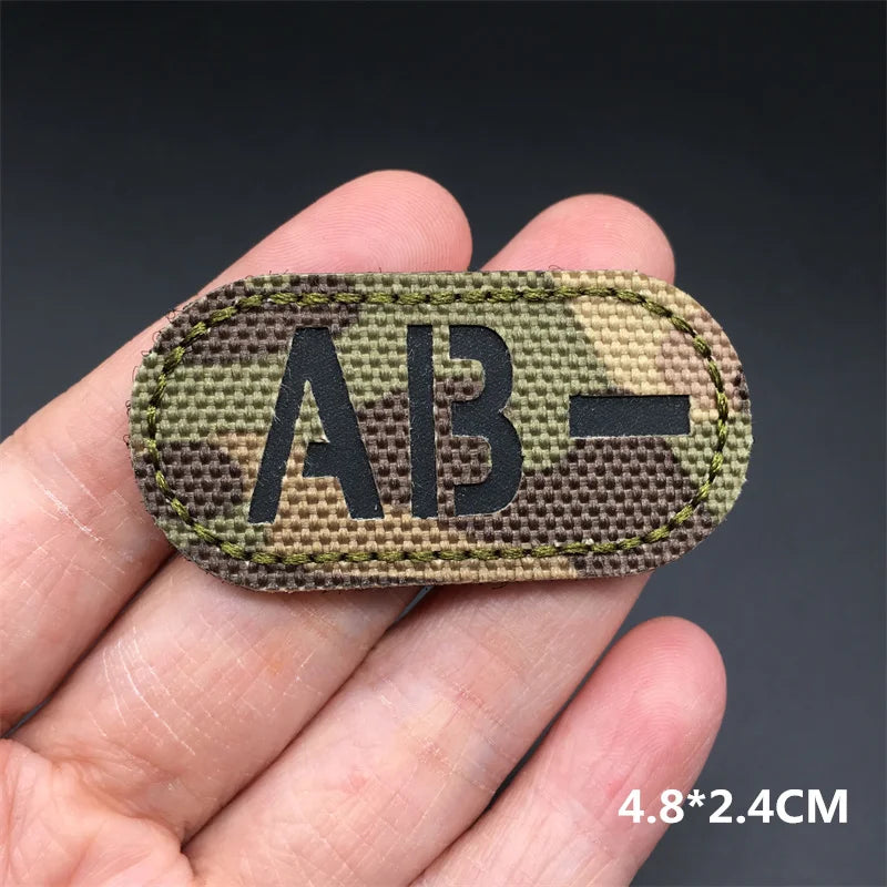 Hand holding an AB- multicam IR reflective tactical blood type hook and loop patch approximately 4.8x2.4cm on a dark background