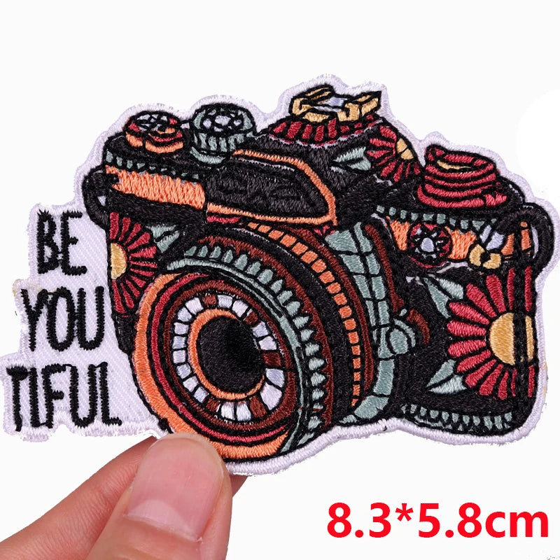 Colorful camera patch with 'Be You Tiful' text held by a hand, on a white background.