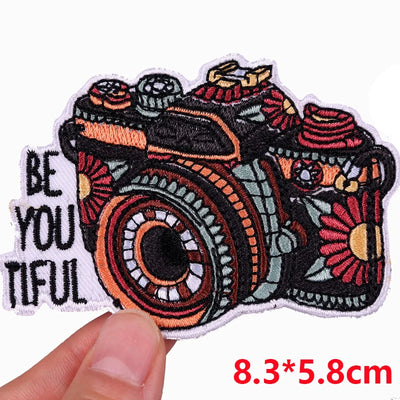 Colorful camera patch with 'Be You Tiful' text held by a hand, on a white background.