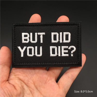 Black patch with white text 'BUT DID YOU DIE?' held in a hand against a dark background