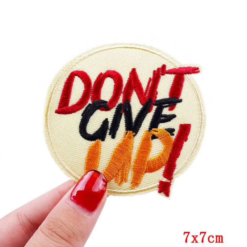 Embroidered patch with 'DON'T GIVE UP!' text held by a hand with red nail polish on a white background.