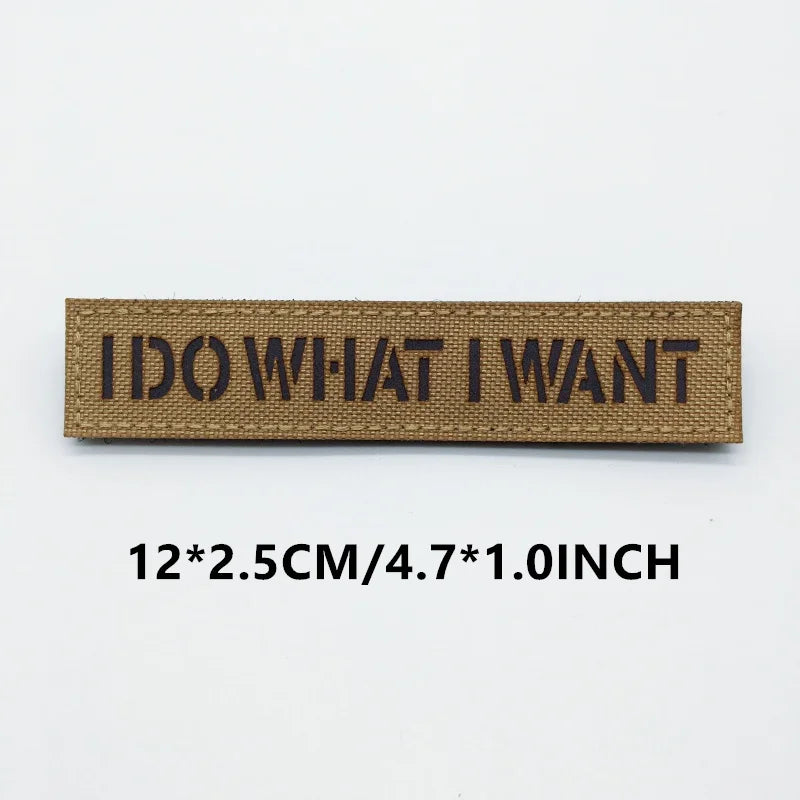 Hand holding a tan and black I Do What I Want IR reflective hook-and-loop morale patch approximately 12x2.5cm on a light background