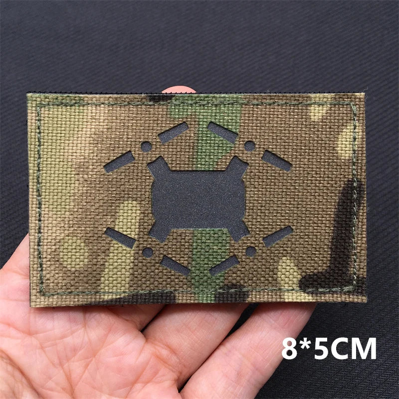 Hand holding a multicam Claymore style IR hook-and-loop tactical badge approximately 8x5cm on a dark background