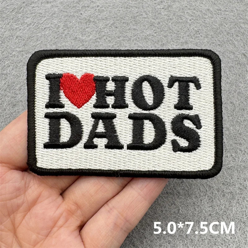 Hand holding a black white and red embroidered I Love Hot Dads iron-on patch approximately 5x7.5cm on a gray background