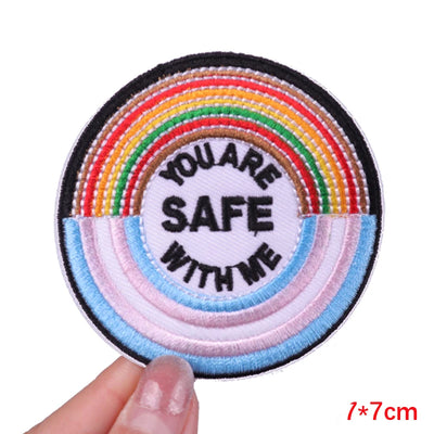 Round patch with rainbow design and 'You are safe with me' text held by a hand.