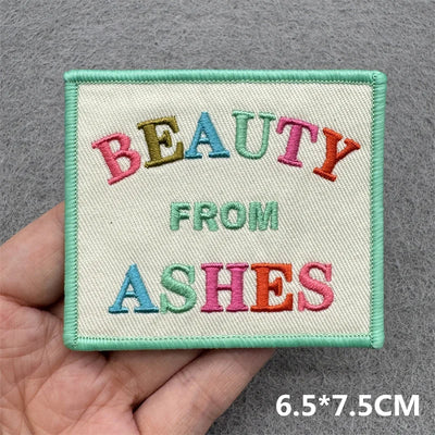 Embroidered patch with 'BEAUTY FROM ASHES' text held by a hand on a gray background