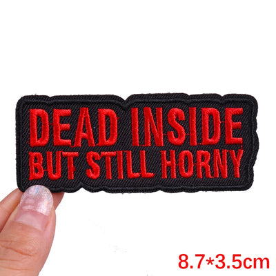 Embroidered patch with red text 'DEAD INSIDE BUT STILL HORNY' held by a hand on a white background.