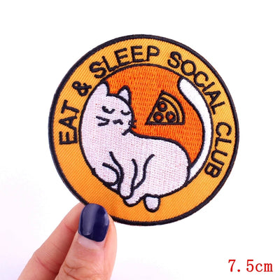 Circular patch with a cat and pizza design, labeled 'Eat & Sleep Social Club', held by a hand.