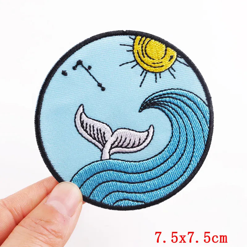 Circular embroidered patch with whale tail, sun, and stars held by a hand on a white background.