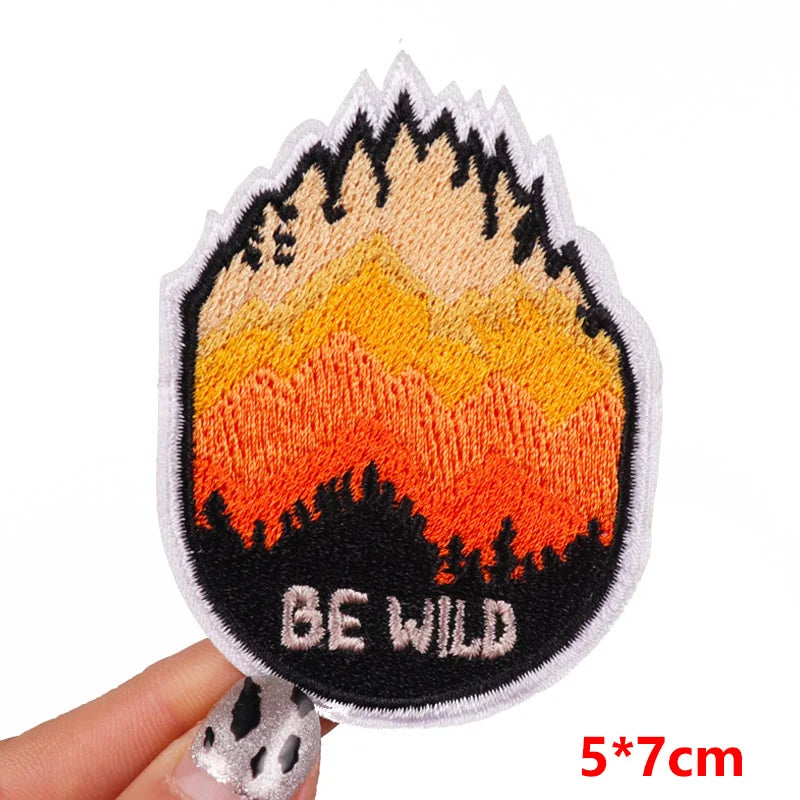Embroidered patch with mountain design and 'BE WILD' text held by a hand on a white background.