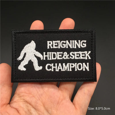 Patch with 'Reigning Hide & Seek Champion' text and a silhouette of a person on a black background, held in a hand.