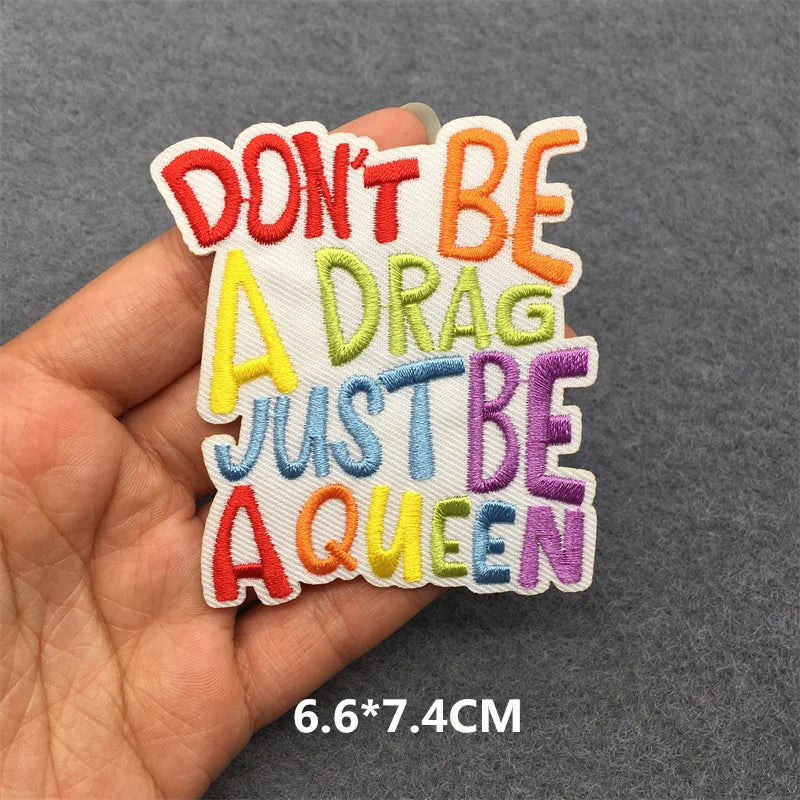 Colorful patch with text 'Don't be a drag, just be a queen' held by a hand on a gray background.